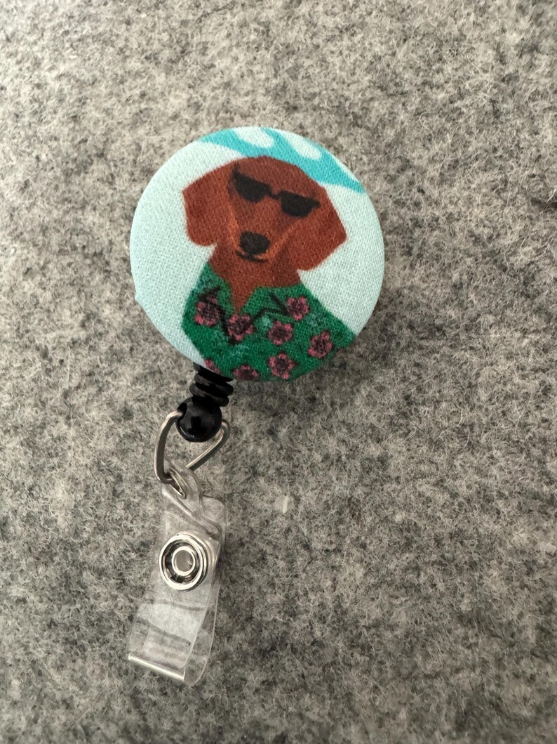 May include: A round retractable badge reel featuring a cartoon dachshund wearing sunglasses and a Hawaiian shirt. The dog is brown with a light blue background and a black retractable cord and clip.