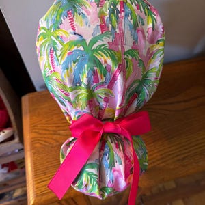 May include: A colorful scrub cap featuring a tropical palm tree pattern in pink, green, blue, and yellow. The cap is tied with a bright pink bow. The cap is made of fabric and is designed to be worn in a medical setting.