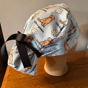 May include: A blue surgical cap with a pattern of golden retrievers and daisies. The cap has a black ribbon tied at the back. The text "Life is golden." is repeated throughout the pattern.