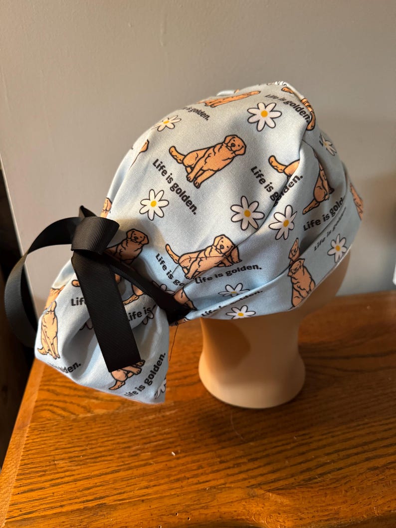 May include: A blue surgical cap with a pattern of golden retrievers and daisies. The cap has a black ribbon tied at the back. The text "Life is golden." is repeated throughout the pattern.