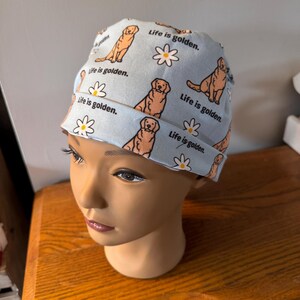 May include: A light blue surgical cap with a pattern of golden retrievers and daisies. The cap has the text "Life is golden." repeated throughout the pattern.