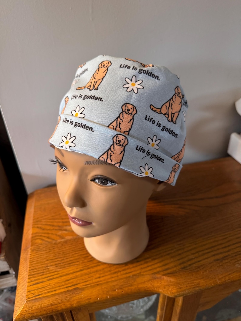 May include: A light blue surgical cap with a pattern of golden retrievers and daisies. The cap has the text "Life is golden." repeated throughout the pattern.