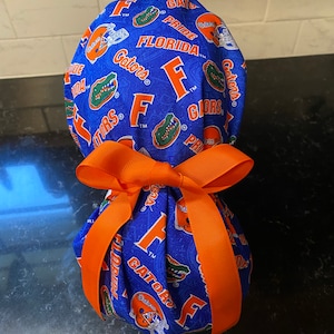 Peut inclure: Un bonnet de chirurgien en tissu bleu et orange avec un nœud. Le bonnet est décoré du logo des Gators de l'Université de Floride et des mots "Gators", "Florida", "Pride" et "F".