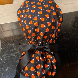 Op de afbeelding: Zwarte scrub-muts van stof met een candy corn-patroon. De muts is vastgebonden met een zwart lint.