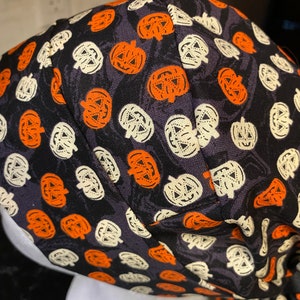 Puede incluir: Un turbante de tela negro con un patr&oacute;n repetido de caras de calabaza naranja y blancas.