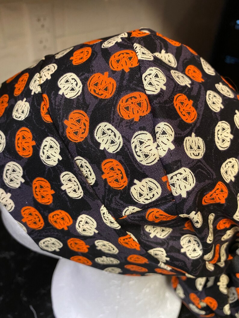 Puede incluir: Un turbante de tela negro con un patr&oacute;n repetido de caras de calabaza naranja y blancas.