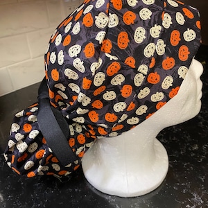 Puede incluir: Un gorro de quir&oacute;fano negro con un patr&oacute;n de calabazas blancas y naranjas. El gorro tiene una corbata negra que se puede atar en la parte posterior de la cabeza.