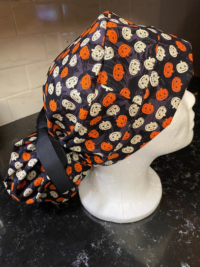 Puede incluir: Un gorro de quir&oacute;fano negro con un patr&oacute;n de calabazas blancas y naranjas. El gorro tiene una corbata negra que se puede atar en la parte posterior de la cabeza.