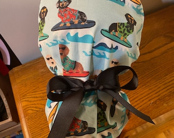 May include: A light blue scrub cap with a repeating pattern of dachshunds surfing on colourful surfboards. The dogs wear sunglasses and Hawaiian shirts. A dark brown ribbon is tied in a bow at the base of the cap.