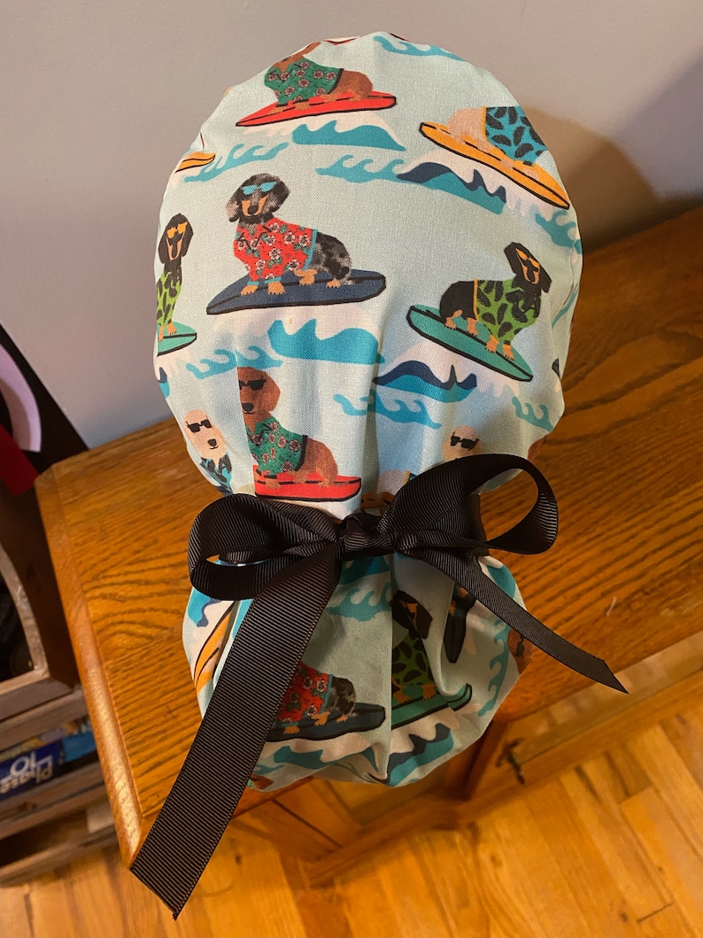 May include: A light blue surgical scrub cap featuring a repeating pattern of dachshunds surfing on colorful surfboards. The dogs wear sunglasses and Hawaiian shirts. A dark brown ribbon is tied in a bow at the base of the cap.