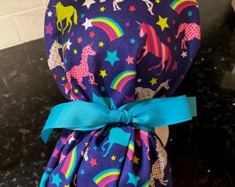 May include: A blue surgical cap with a unicorn, rainbow, and star pattern. The cap is tied with a turquoise ribbon.
