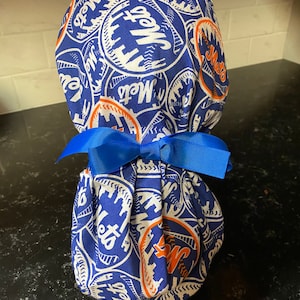 May include: Blue and white fabric scrub hat with a repeating pattern of baseballs with the word "Mets" on them. The hat has a blue ribbon tied around it.