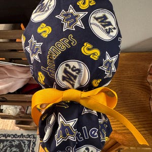 Peut inclure: Un bonnet de chirurgien bleu marine avec un motif de balles de baseball blanches, d'étoiles jaunes et le logo des Mariners de Seattle. Le bonnet est noué avec un ruban jaune vif. Le mot "Mariners" est inclus.