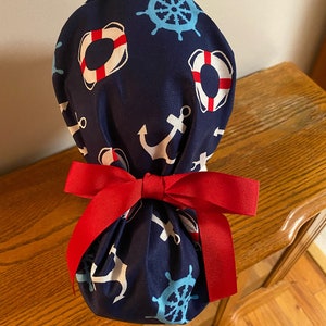 May include: A blue fabric scrub hat with a nautical theme. The hat is decorated with anchors, life preservers, and ship wheels. It is tied with a red ribbon.