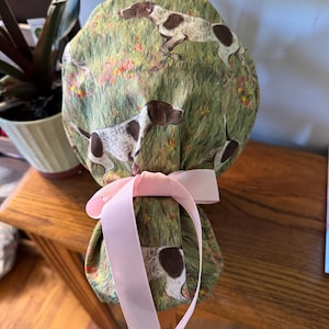 May include: A green and white patterned scrub cap featuring brown and white dogs in a field of flowers. The cap is tied with a light pink ribbon. The cap is made of fabric.