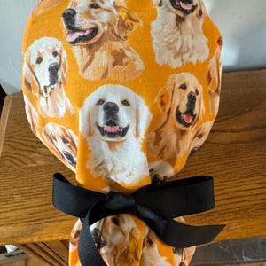 May include: A yellow fabric scrub hat with a black ribbon tied in a bow. The fabric features a repeating pattern of golden retriever dog faces.