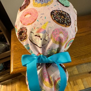 May include: A pink and white fabric scrub hat with a donut and unicorn print. The hat is tied with a bright blue ribbon.