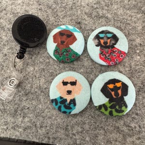 May include: A black retractable badge reel next to four round pinback buttons. Each button features a cartoon dachshund wearing sunglasses and a Hawaiian shirt. The dachshunds are brown, black, and tan, with light blue backgrounds.