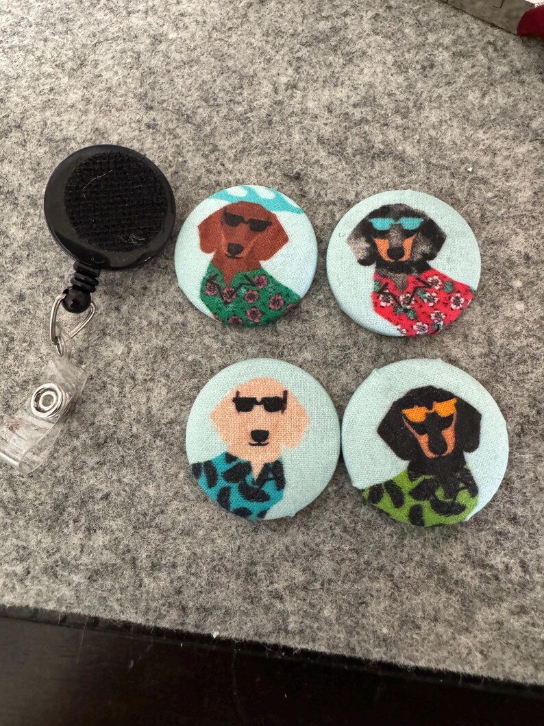 May include: A black retractable badge reel next to four round pinback buttons. Each button features a cartoon dachshund wearing sunglasses and a Hawaiian shirt. The dachshunds are brown, black, and tan, with light blue backgrounds.