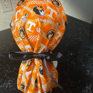 May include: A patterned orange and white scrub cap featuring the word "Tennessee", the letter "T", and the word "VOLS". The cap has a black ribbon tied in a bow. The cap is made of fabric.