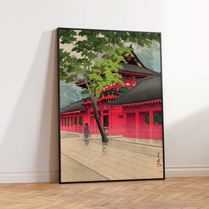 May include: A traditional Japanese woodblock print depicting a red temple with a green tree in the foreground. The temple has a black roof and white walls with red accents. The print is framed in black.