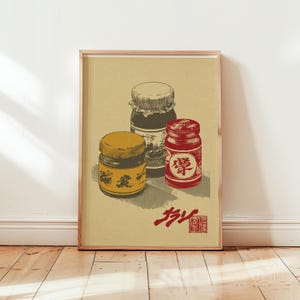 Op de afbeelding: Een ingelijste print met drie vintage potten met etiketten en deksels. De potten zijn in tinten geel, rood en bruin, tegen een beige achtergrond. Japanse tekst is onderaan zichtbaar. Het kunstwerk wordt op een houten vloer getoond.