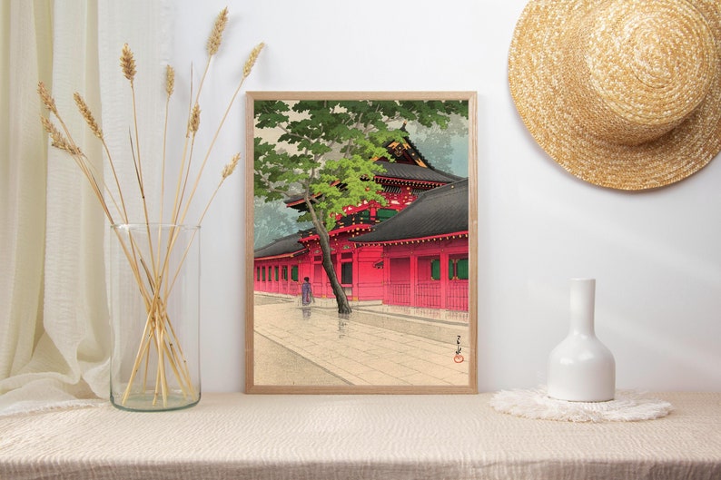 May include: A framed print of a traditional Japanese woodblock print depicting a red temple with a green tree in the foreground. The print is set against a white wall with a straw hat and a white vase on a shelf.