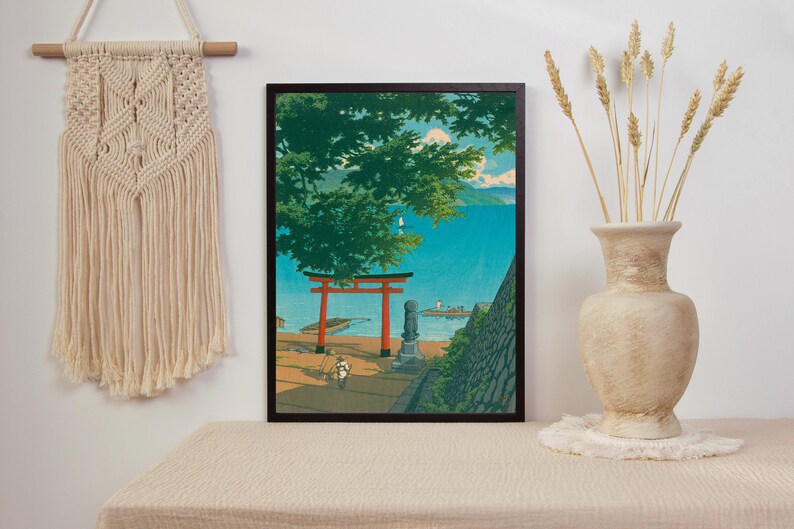 May include: A framed print of a Japanese landscape with a red torii gate, a stone statue, and a view of a blue body of water. The print is set against a white wall with a macrame wall hanging and a vase of dried wheat.