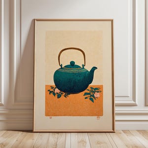 May include: Framed art print of a teal teapot with a brown handle and gold detailing. The teapot is set on an orange surface with floral accents. The background is a light beige colour.