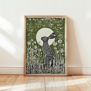 May include: A framed print featuring a black and white illustration of a hare in a field of flowers, with a full moon in the background. The artwork has a rustic, woodcut style, with a green and white color palette. The print is displayed on a wooden floor.