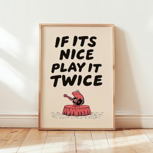 Op de afbeelding: Een ingelijste kunstdruk op een lichtcrème achtergrond met de zwarte tekst IF ITS NICE PLAY IT TWICE. Onder de tekst staat een rode illustratie van een kikker die op een boomstronk zit en op een zwarte akoestische gitaar speelt.