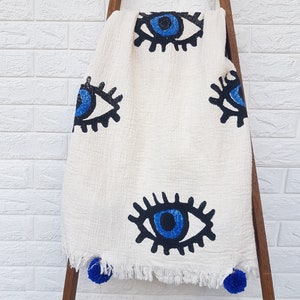 May include: White cotton throw blanket with a fringe border and blue evil eye designs. The blanket has blue pom-poms on the corners.