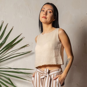 May include: A woman wearing a cream sleeveless top with a slightly cropped hem and a pair of striped trousers with a brown belt.