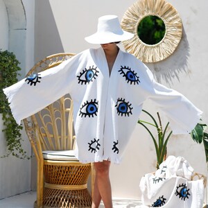 Op de afbeelding: Een witte kimono-stijl badjas met een herhalend, handgeschilderd grafisch patroon van blauwe en zwarte ogen. Het kledingstuk heeft wijde, losse mouwen, een open voorkant en een gefranjerde zoom. Een bijpassend wit stuk stof is ook zichtbaar.