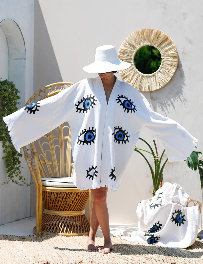 Op de afbeelding: Een witte kimono-stijl badjas met een herhalend, handgeschilderd grafisch patroon van blauwe en zwarte ogen. Het kledingstuk heeft wijde, losse mouwen, een open voorkant en een gefranjerde zoom. Een bijpassend wit stuk stof is ook zichtbaar.