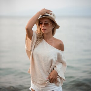 May include: A beige off-the-shoulder linen top with wide sleeves, paired with a straw hat featuring a black band and rectangular tinted sunglasses with thin gold frames. The outfit is completed by white denim shorts with a distressed hemline.