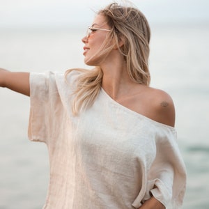 May include: A beige, short-sleeved, off-the-shoulder top with a loose, boxy fit and a visible woven texture. The garment features a wide neckline that rests off one shoulder, revealing a small, black, eye-shaped tattoo on the upper arm area.