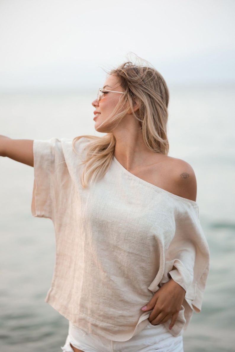 May include: A beige, short-sleeved, off-the-shoulder top with a loose, boxy fit and a visible woven texture. The garment features a wide neckline that rests off one shoulder, revealing a small, black, eye-shaped tattoo on the upper arm area.