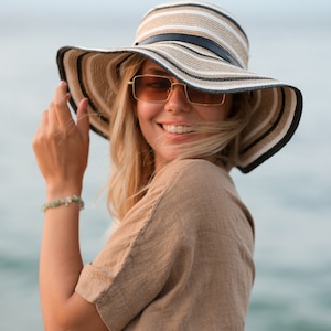 May include: A wide-brimmed straw hat featuring horizontal stripes in shades of beige, white, and black, accented with a thin black band around the crown. The hat is paired with rectangular sunglasses with gold-toned frames and amber-tinted lenses.