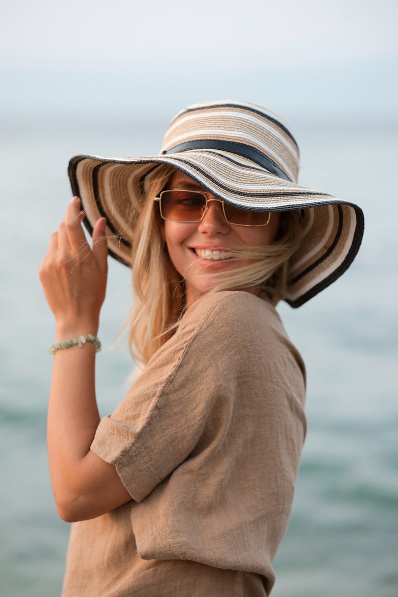 May include: A wide-brimmed straw hat featuring horizontal stripes in shades of beige, white, and black, accented with a thin black band around the crown. The hat is paired with rectangular sunglasses with gold-toned frames and amber-tinted lenses.