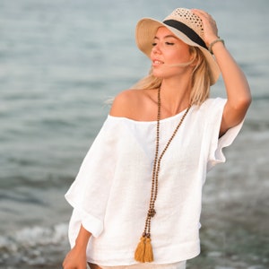 May include: A beige straw hat with a black ribbon band, a loose-fitting white short-sleeved top, and a long necklace featuring small brown wooden beads and two mustard yellow fabric tassels hanging down the front of the garment.