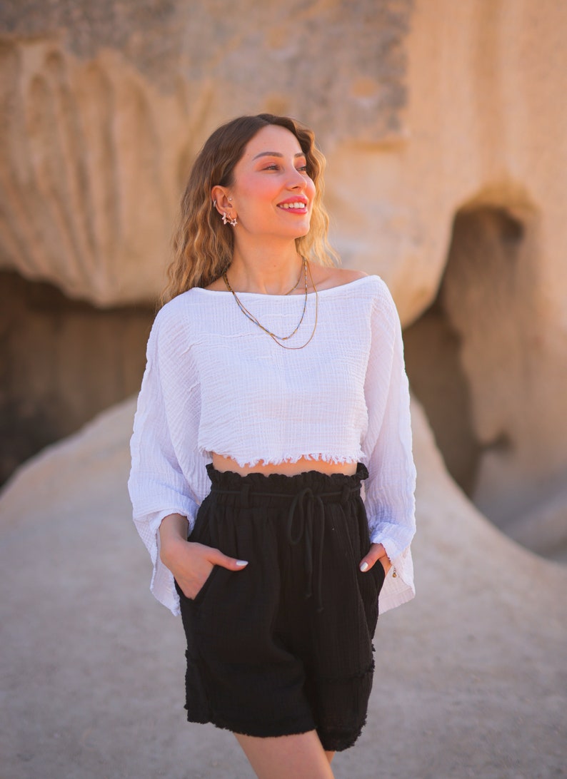 May include: A woman wearing a white, off-the-shoulder, cropped top with a frayed hem and black, high-waisted shorts with a drawstring. She is standing in front of a rocky, desert-like landscape.