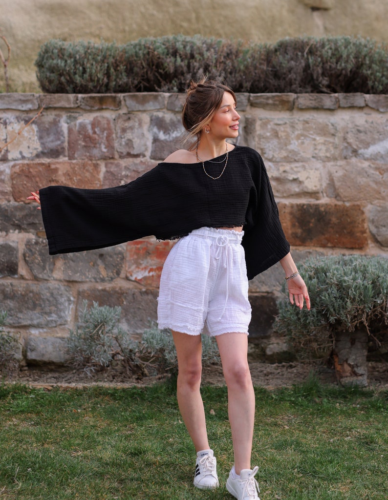 May include: A woman wearing a black, cropped, off-the-shoulder top with white, ruffled shorts and white sneakers.