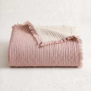 May include: A folded rectangular throw blanket featuring a textured, crinkled fabric surface. The top side is a dusty rose pink, while the underside is a contrasting cream color. The edges are finished with a decorative, frayed fringe trim.