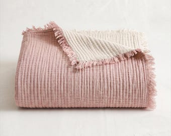 Puede incluir: Una manta rectangular doblada con una superficie de tela texturizada y arrugada. El lado superior es de color rosa empolvado, mientras que el reverso es de color crema. Los bordes de la manta están acabados con un fleco decorativo deshilachado.