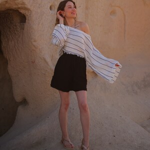 Puede incluir: Una blusa blanca de manga larga con hombros descubiertos, finas rayas horizontales negras y dobladillo deshilachado, combinada con pantalones cortos negros con cintura de cord&oacute;n y bordes deshilachados, y sandalias beige de tiras con suela plana.