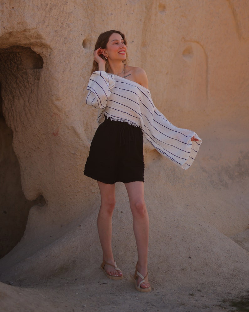 Puede incluir: Una blusa blanca de manga larga con hombros descubiertos, finas rayas horizontales negras y dobladillo deshilachado, combinada con pantalones cortos negros con cintura de cord&oacute;n y bordes deshilachados, y sandalias beige de tiras con suela plana.