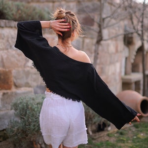 May include: A woman wearing a black, off-the-shoulder, cropped top with long sleeves and white, high-waisted shorts. The top has a frayed hemline. The woman is standing in front of a stone wall with a potted plant in the background.