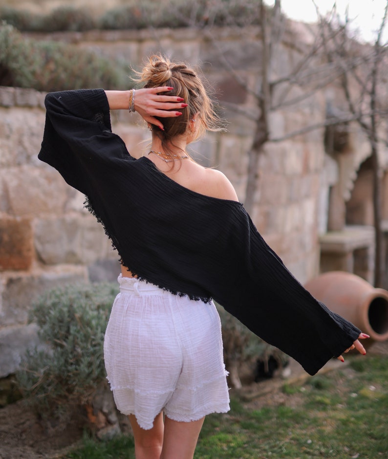May include: A woman wearing a black, off-the-shoulder, cropped top with long sleeves and white, high-waisted shorts. The top has a frayed hemline. The woman is standing in front of a stone wall with a potted plant in the background.