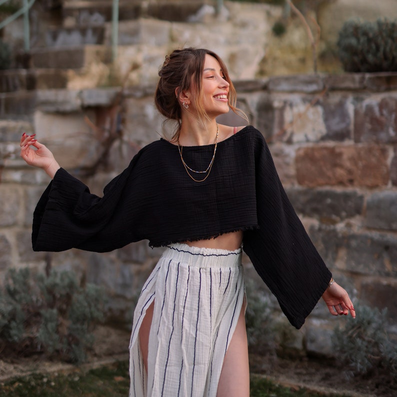 May include: A woman wearing a black cropped top with long sleeves and a white and black striped skirt. She is standing in front of a stone wall and has a gold necklace on.